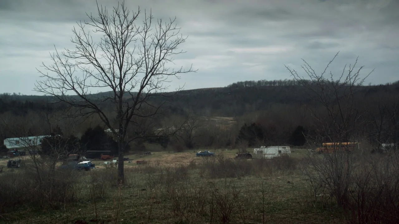 Winter’s Bone (2010) movie still: wide — A desolate rural field under a heavy, overcast grey sky features bare trees. Dilapidated mobile homes and…