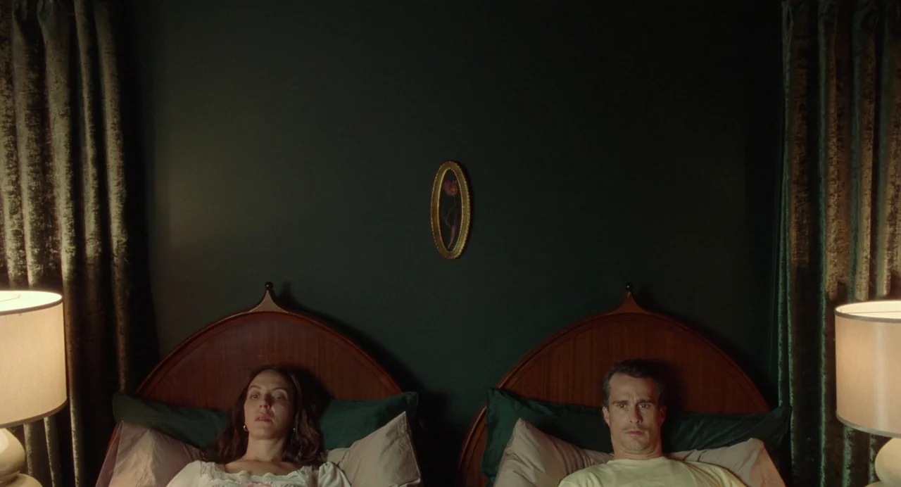 Babysitter (2022) movie still: medium — A symmetric bedroom with dark green walls and a central oval framed artwork. A man and woman lie in separate…