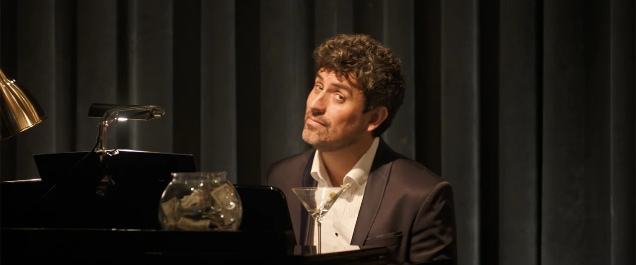 Drop (2025) movie still: medium — A dark lounge with heavy curtains behind a man playing a grand piano. The man sits at the keys while a martini…