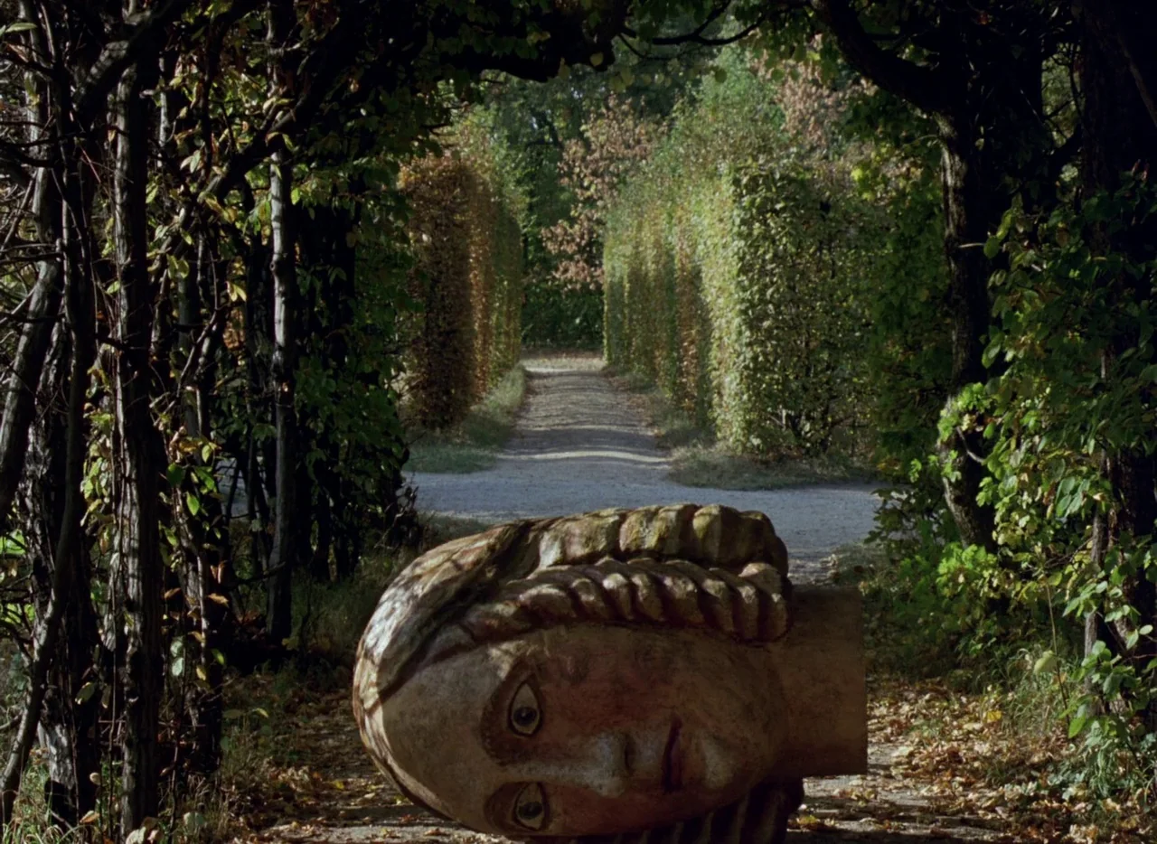 Faust (1994) movie still: wide — A tree-lined pathway with clipped hedges leads into a park at eye level. A large, sculpted stone head sits on the…