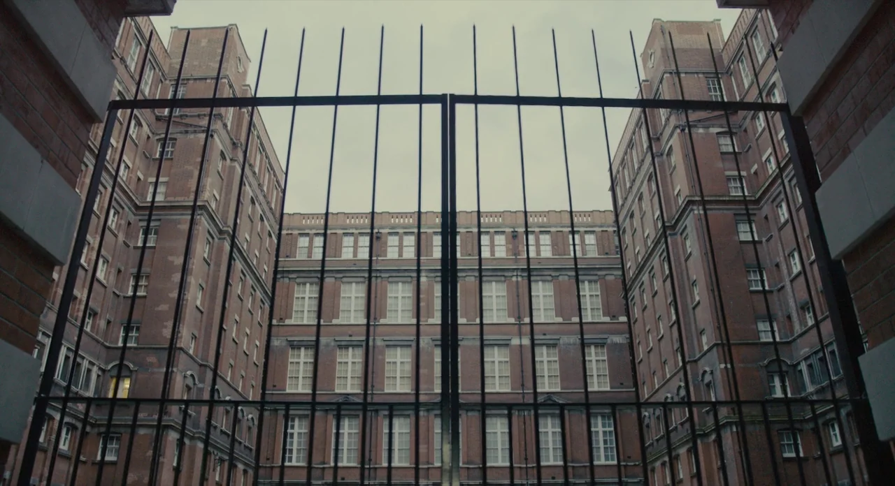 The Danish Girl (2015) movie still: establishing — A U-shaped multi-story red brick apartment complex is viewed through a tall iron gate. The…