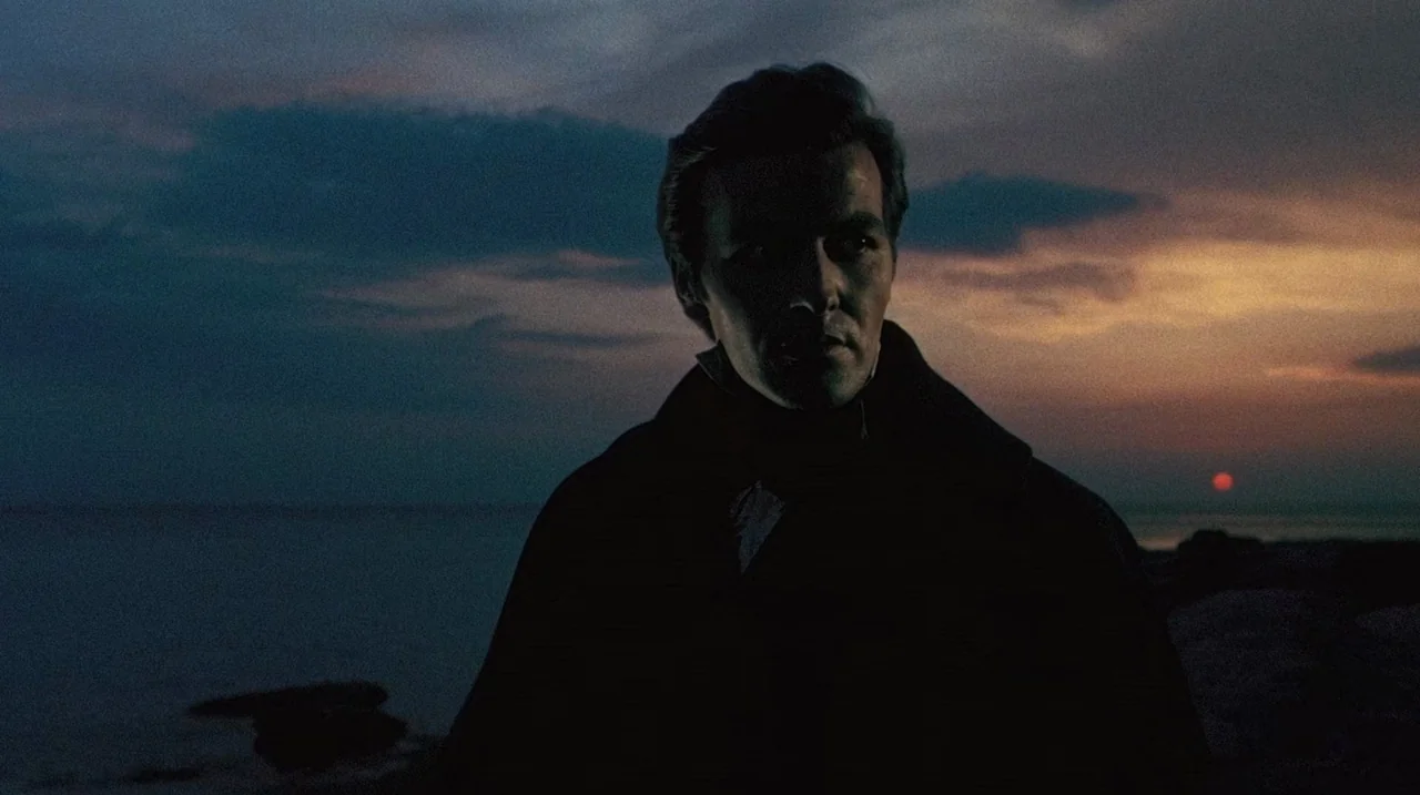 The Whip and The Body (1963) movie still: medium — A man stands before a dark body of water during a twilight sunset. The horizon is glowing with…