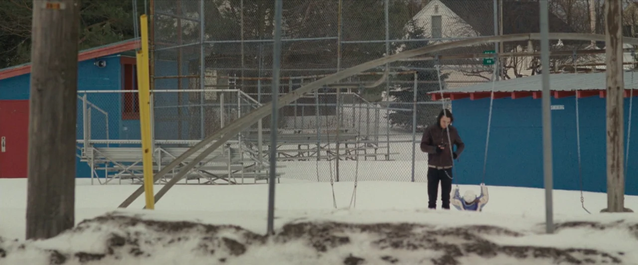 For Ellen (2012) movie still: wide — A snow-covered playground features a blue utility building and metal swing set frames. A man wearing a leather…