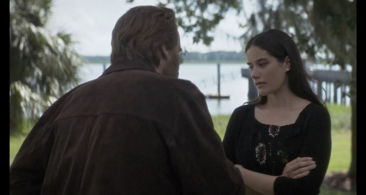 The Poison Rose (2019) movie still: medium — A man and a woman stand near a body of water with trees nearby. They appear to be engaged in…