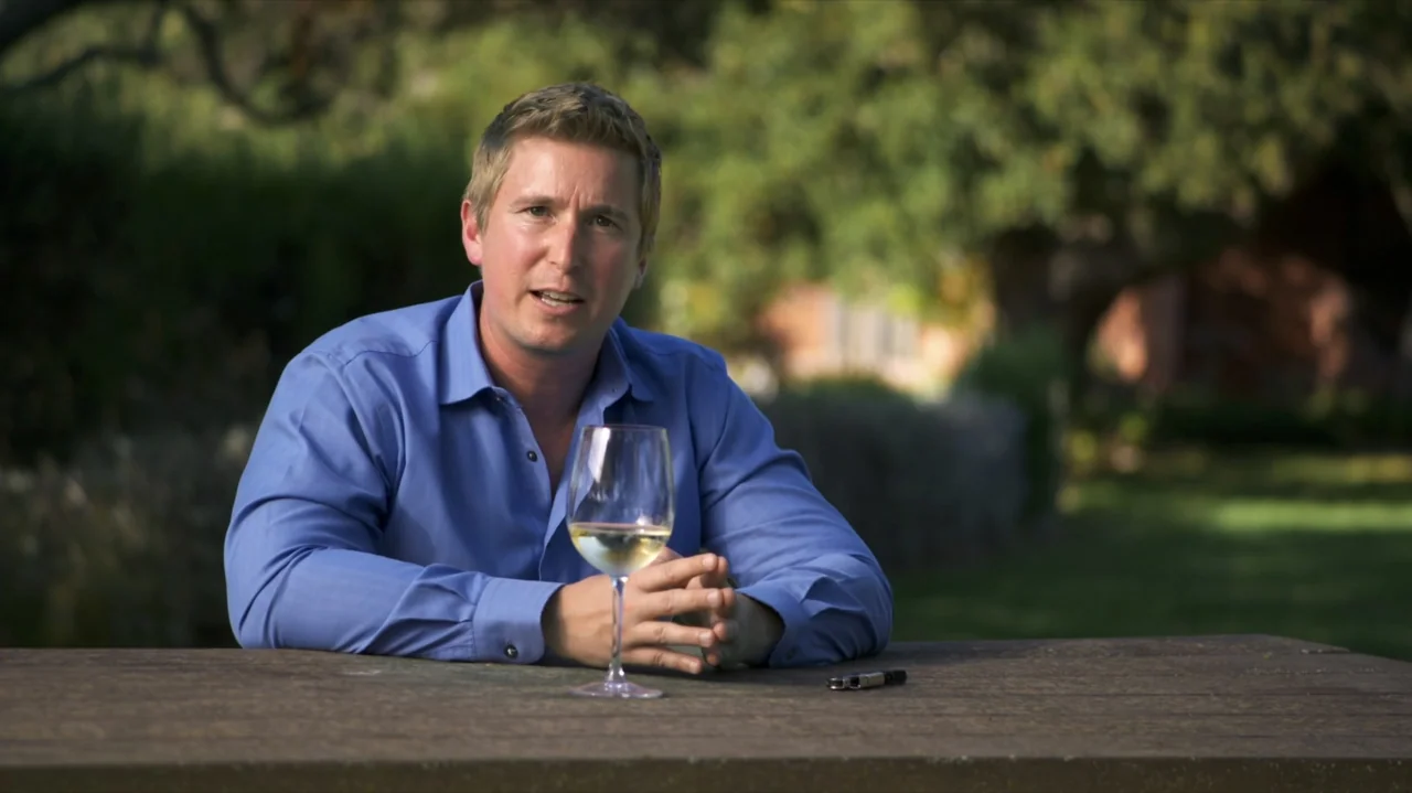 Somm: Into the Bottle (2015) movie still: medium — A man sits at a wooden table in an outdoor park setting during daytime. He rests his hands near a…