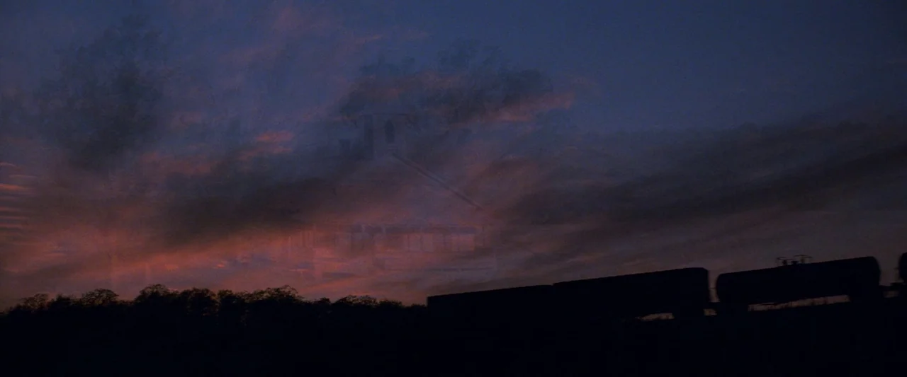 The Outsiders (1983) movie still: establishing — A dark, silhouetted freight train moves across a field during a purple twilight. A ghostly,…