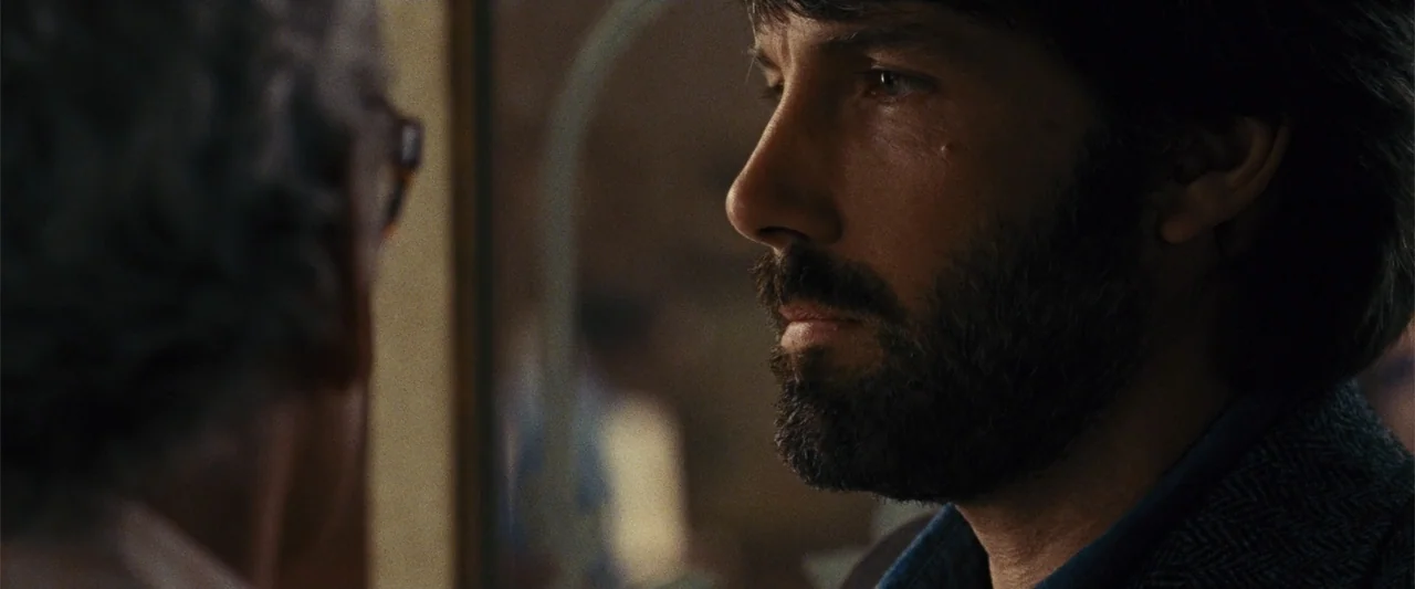 Argo (2012) movie still: closeup — An office interior features a blurred figure in the foreground and a man profile. The man looks toward the unseen…