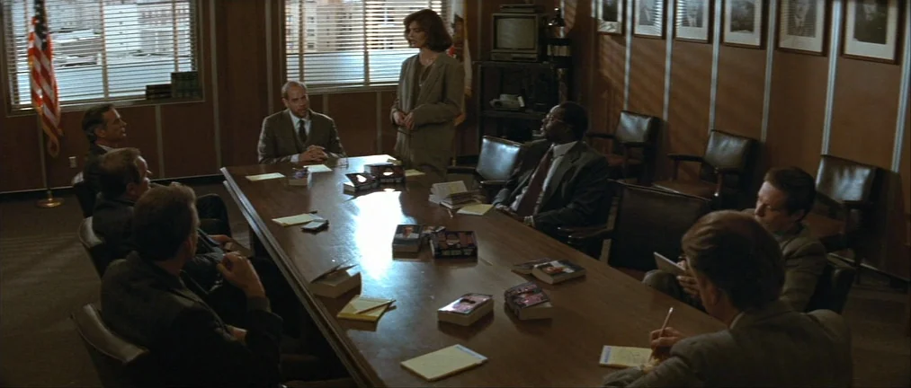 Basic Instinct (1992) movie still: wide — A wood-paneled conference room features a large oval table surrounded by men in suits. A woman stands at…