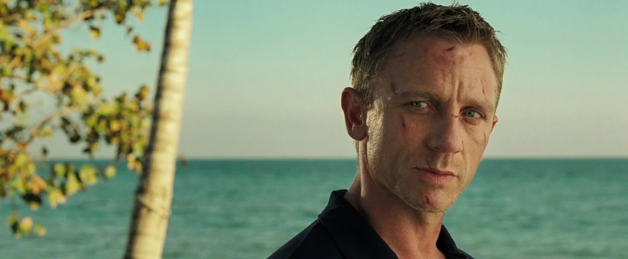 Casino Royale (2006) movie still: closeup — A man stands outdoors near the shoreline of a bright turquoise sea during daytime. The man has small…
