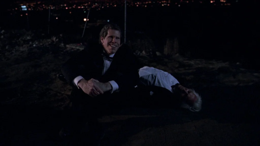 Cronos (1993) movie still: medium — A dark, uneven dirt patch overlooks a blurred city grid at night. One man wearing a tuxedo sits upright smiling…