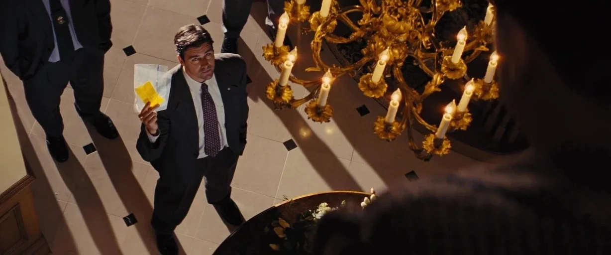 The Wolf of Wall Street (2013) movie still: wide — A man in a suit stands on a patterned tile floor beneath a chandelier. He holds a yellow item…