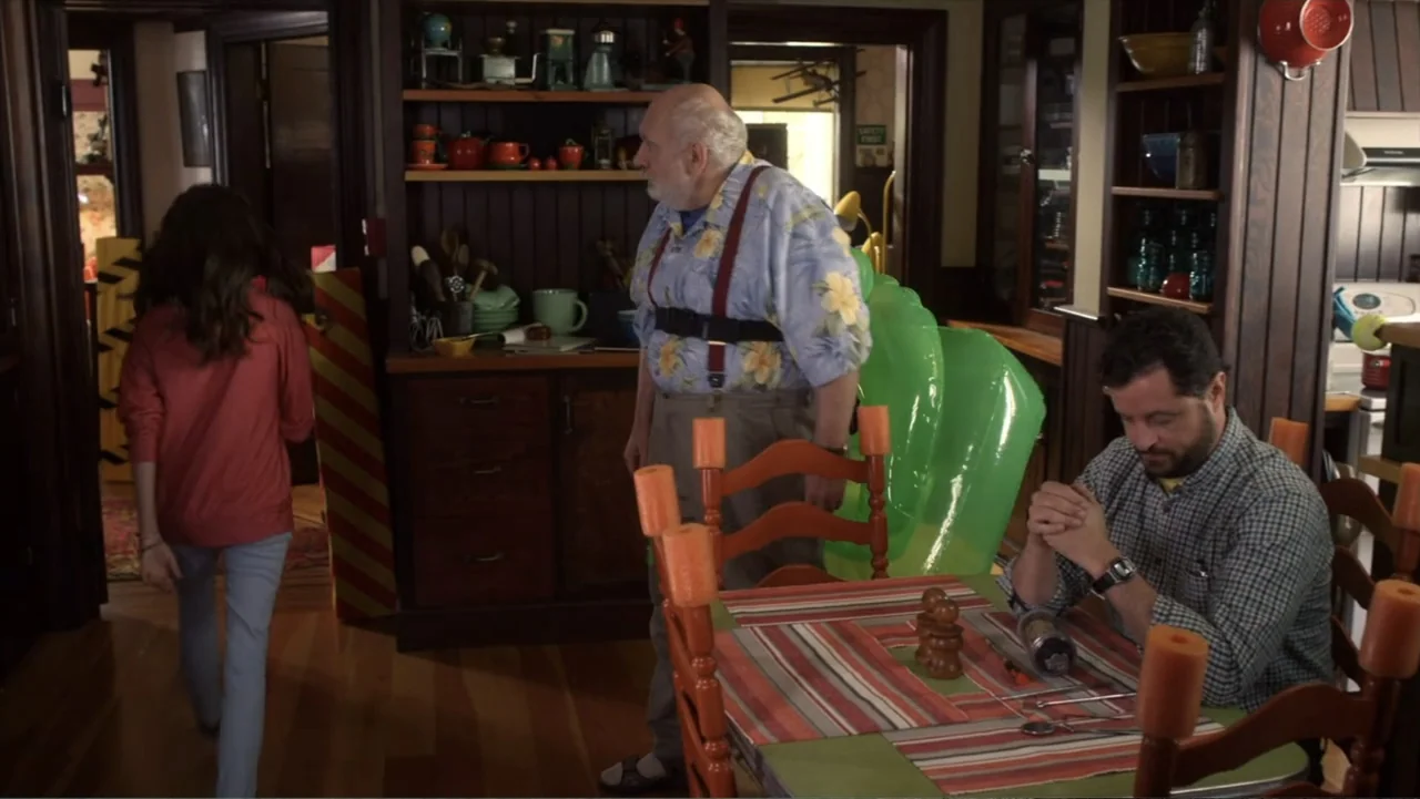 Jinxed (2013) movie still: medium — A residential room features dark wood cabinetry stocked with ceramic dishware and a dining table. A man stands…