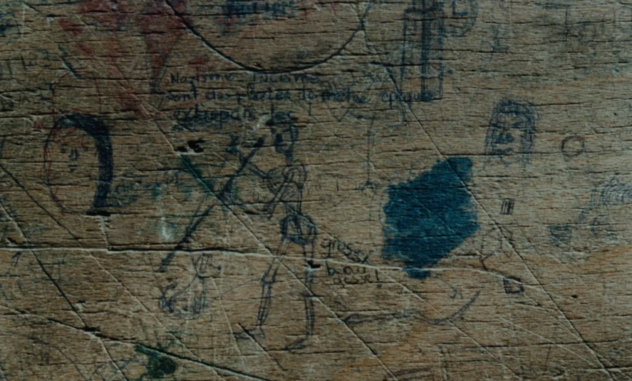 Graduate First (1978) movie still: closeup — A weathered wooden surface is covered in various ink drawings and scratched text markings. Multiple…