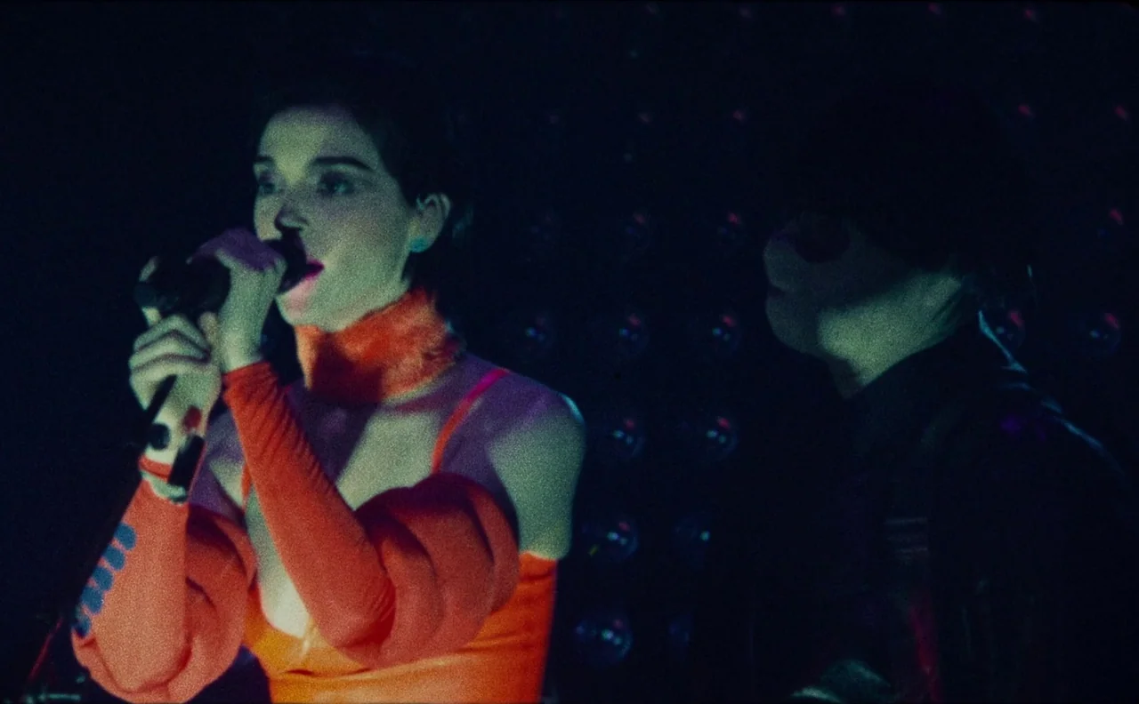 The Nowhere Inn (2020) movie still: medium — A dark concert stage with a repetitive pattern of dim recessed lights behind performers. A woman in…