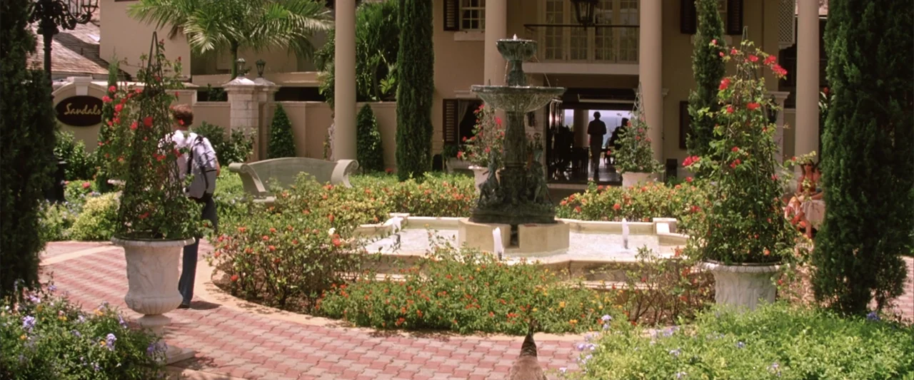 License to Wed (2007) movie still: wide — A resort courtyard features a central stone fountain surrounded by manicured flower beds. A person…