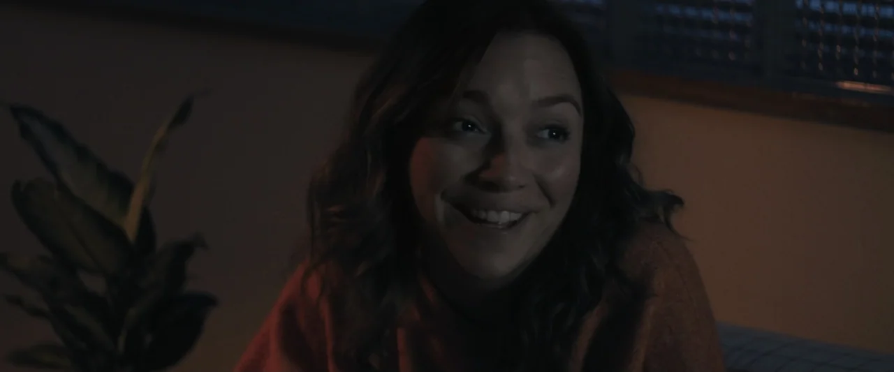 Sign Here (2017) movie still: closeup — A dimly lit room interior features a houseplant in the foreground next to a person. A woman with long wavy…