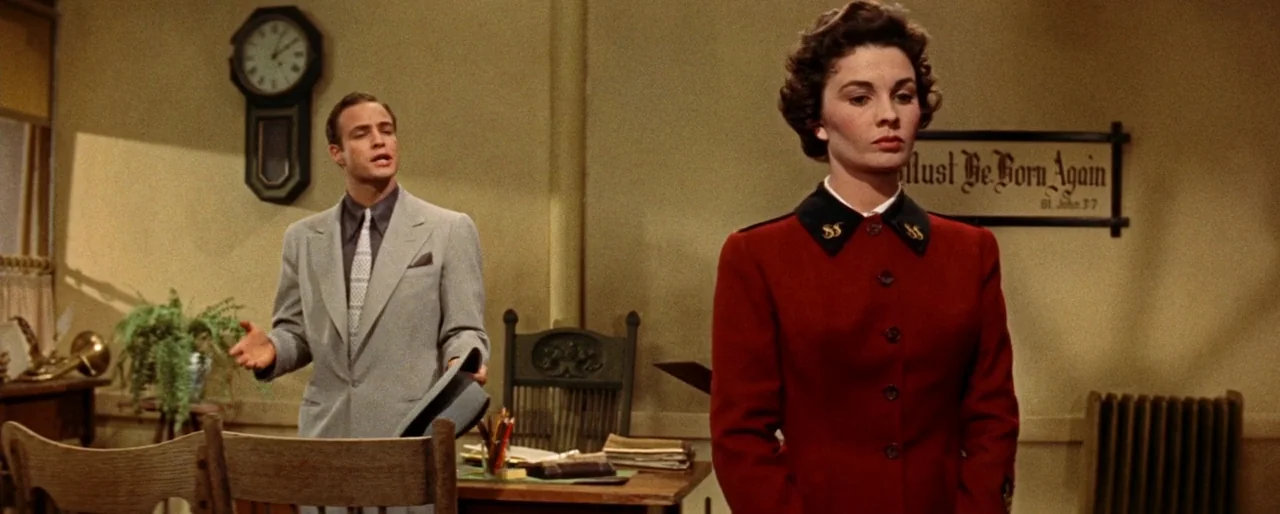 Guys and Dolls (1955) movie still: medium — A modest office space features plain walls adorned with a wall clock and text. A man stands near a…