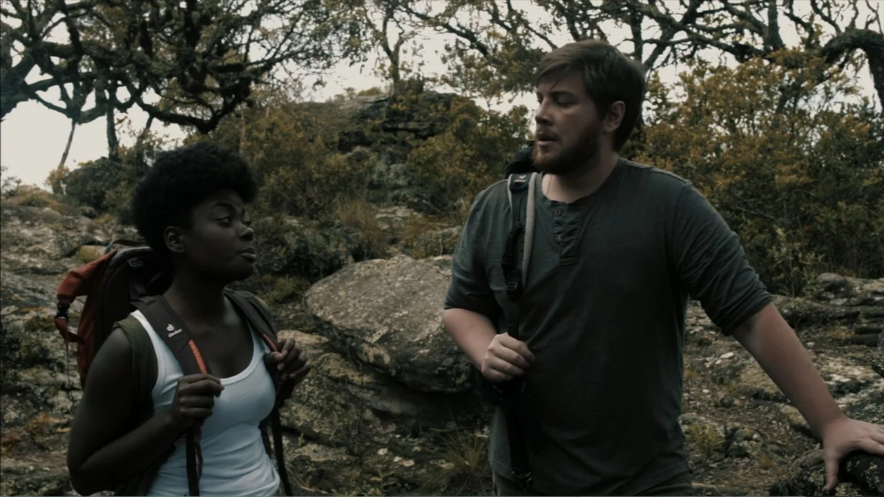 The Grotto (2022) movie still: medium — A rugged outdoor trail features large boulders and scattered trees against an overcast sky. Two hikers with…