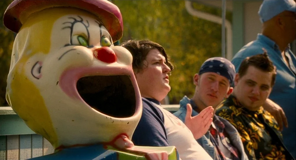 Adventureland (2009) movie still: medium — An outdoor amusement park area features a weathered yellow plastic clown sculpture in foreground. Three…