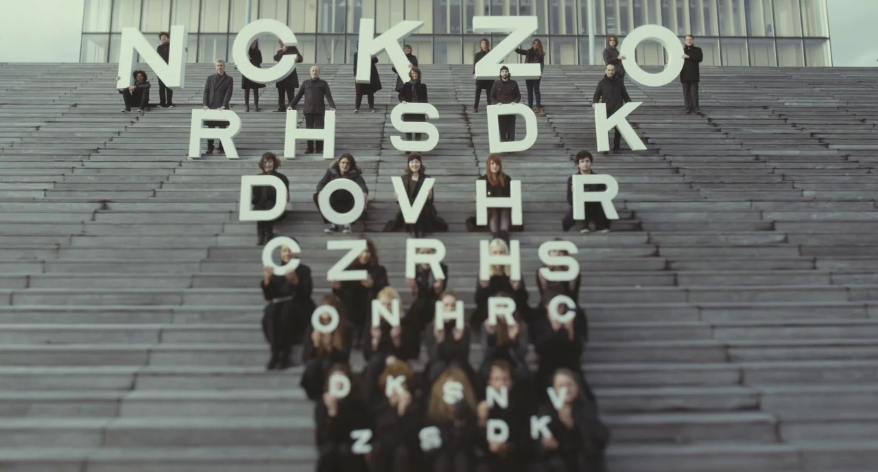 Faces Places (2017) movie still: wide — A group of people stands and sits on broad outdoor concrete stairs. Each individual holds a large white…