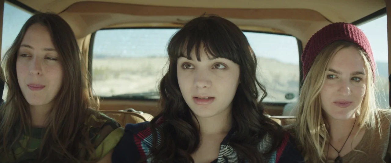 Southbound (2015) movie still: medium — Three young women sit closely together in the backseat of an old vehicle. Outside the rear windshield, a…