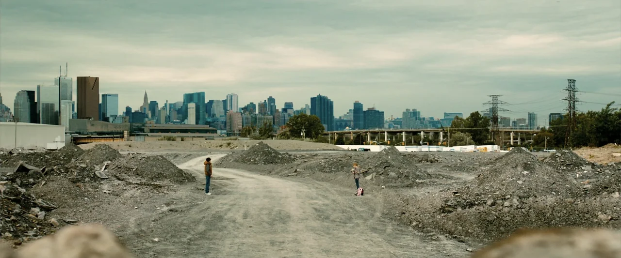 Kick-Ass 2 (2013) movie still: wide — A vast, empty dirt lot filled with piles of rubble lies before a skyline. Two teenagers stand separated on a…