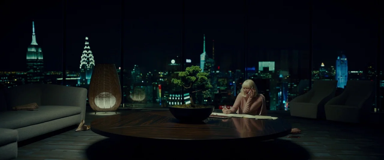 Okja (2017) movie still: wide — A high-rise penthouse office overlooks a brightly lit city skyline at night through windows. An older woman sits on…