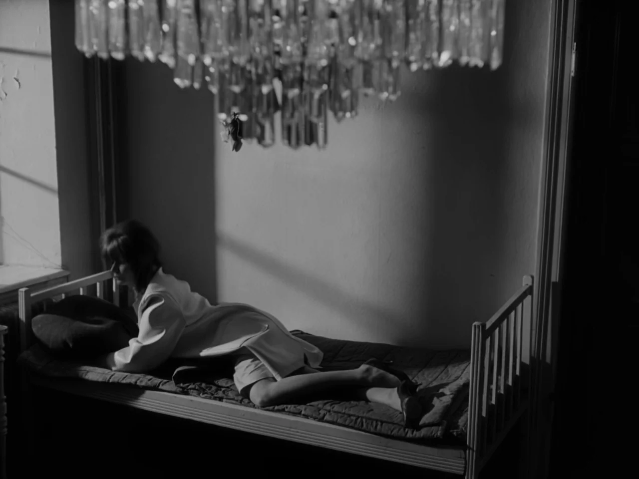 The Baby Carriage (1963) movie still: medium — A spartan room features a single bed and a large out-of-focus crystal chandelier above. A young woman…