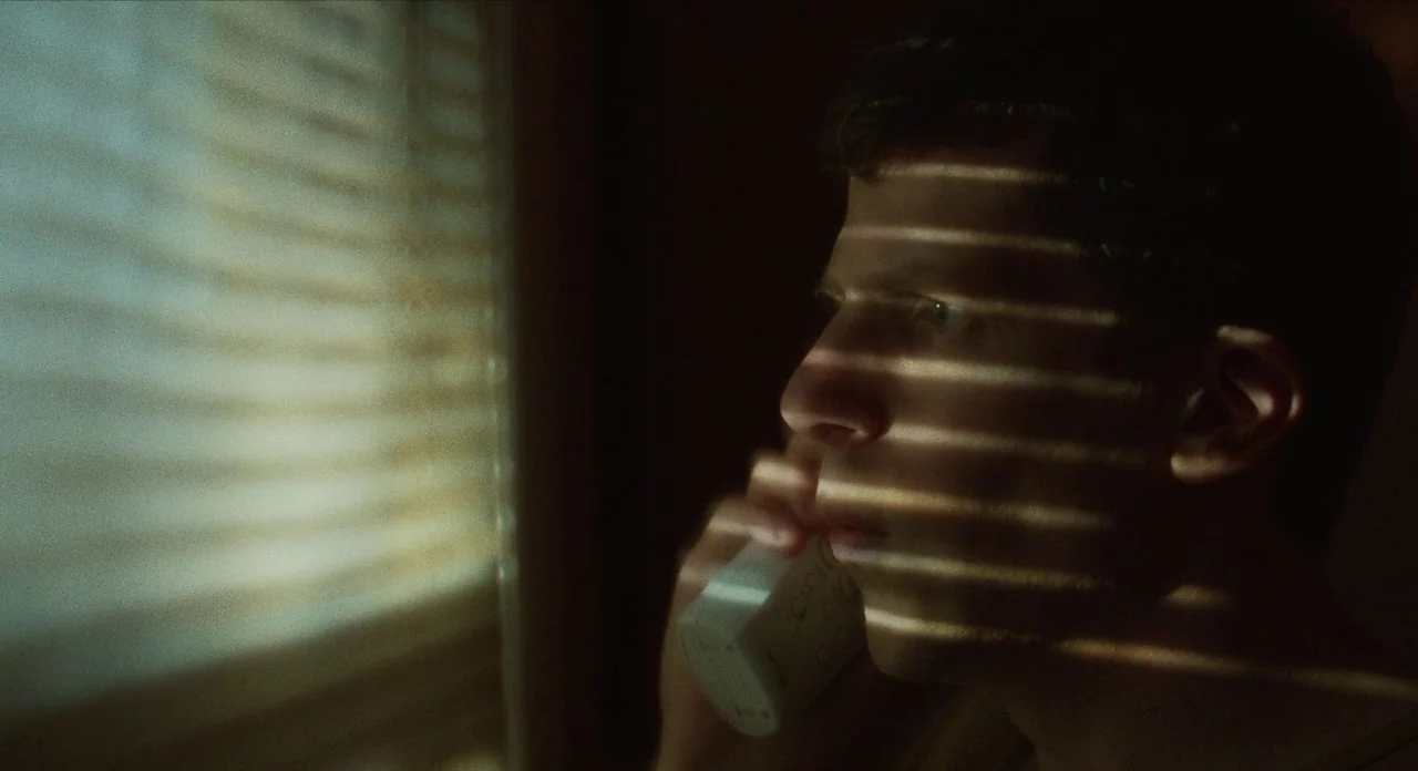 The Art of Self Defence (2019) movie still: closeup — A dark interior room features light filtering through horizontal window blinds. A young man…