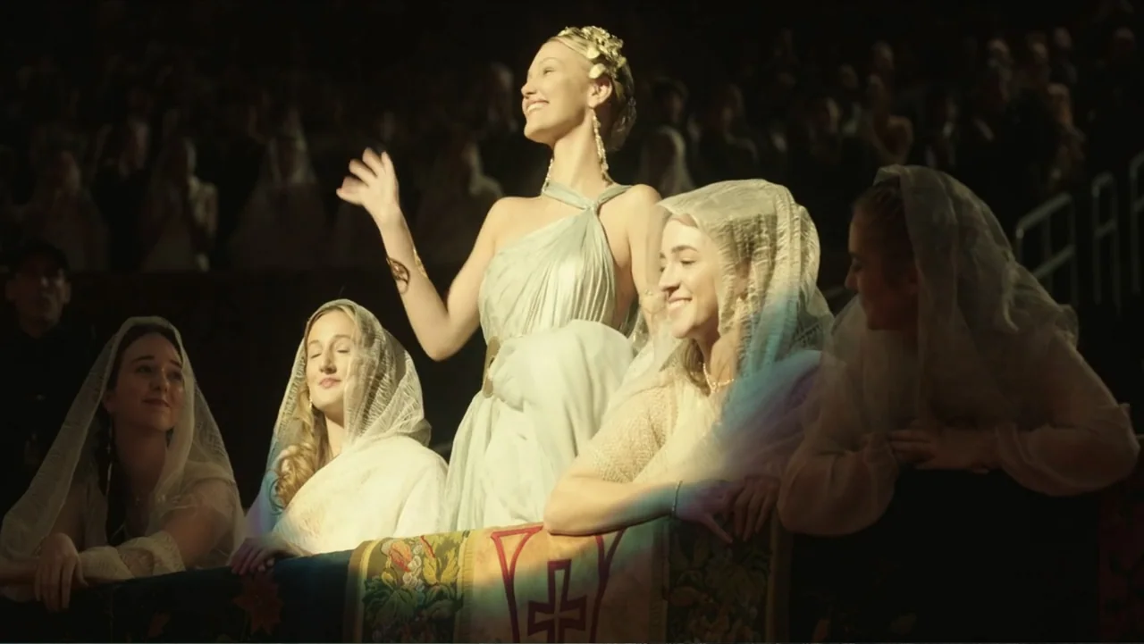 Megalopolis (2024) movie still: medium — A group of women in formal attire sit inside a dimly lit theater box. The women are dressed in…