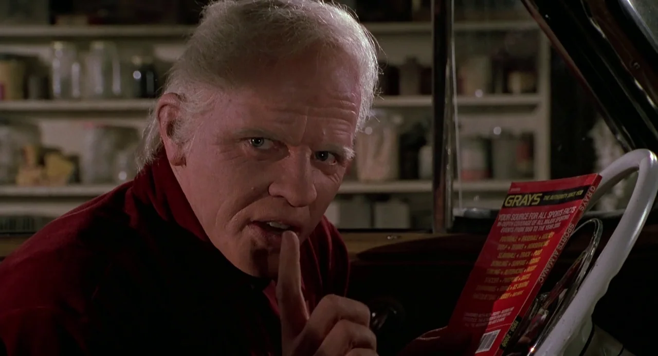 Back to the Future II (1989) movie still: closeup — An older man sits inside a vehicle at night holding a sports almanac. He holds a finger to his…