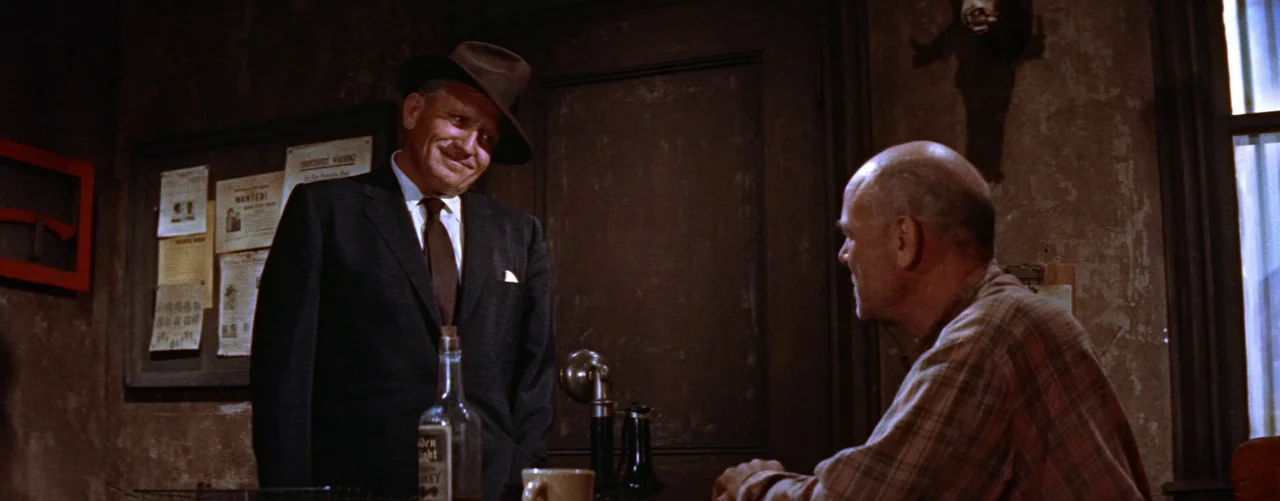 Bad Day At Black Rock (1955) movie still: medium — A dimly lit office features textured walls decorated with wanted posters and notices. A man…