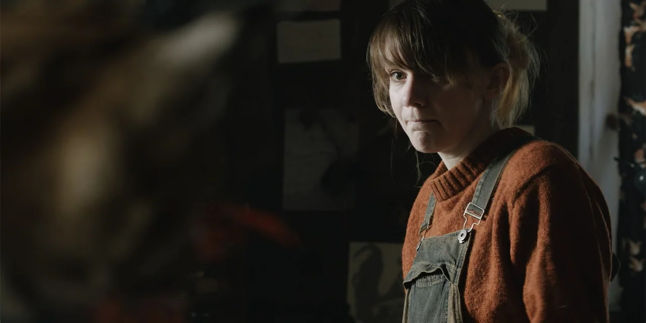 La coda del diavolo (2024) movie still: medium — A dark, cluttered interior workspace contains papers and equipment on a wall. A young woman in a…