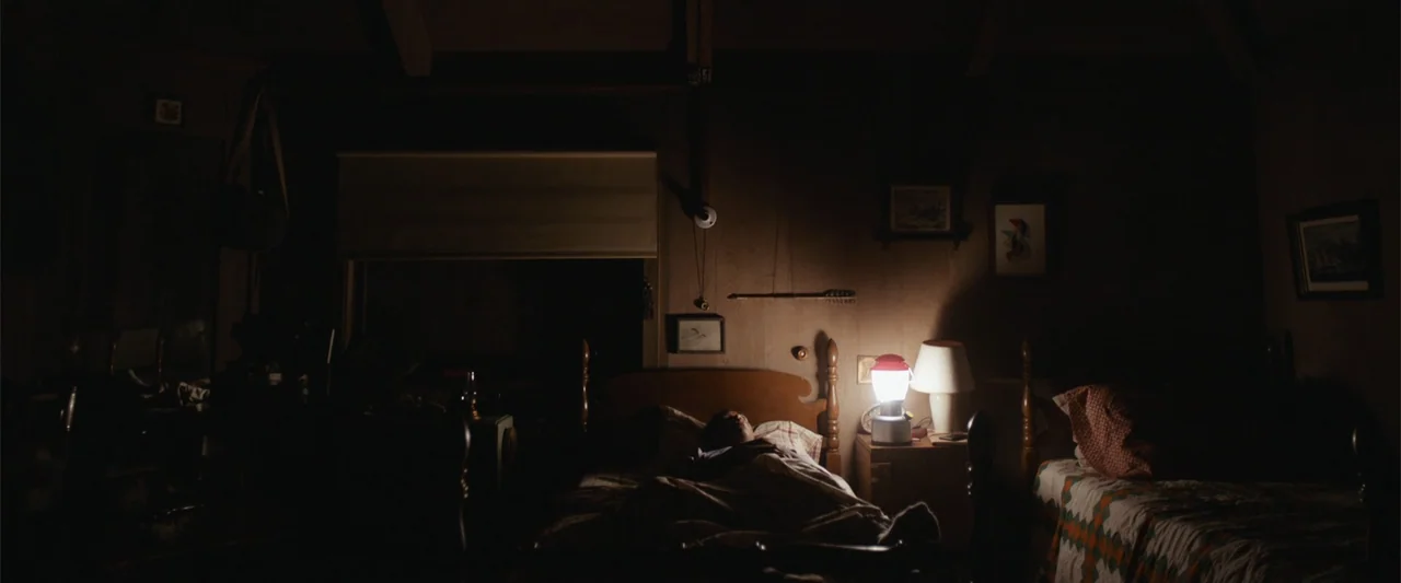 It Comes at Night (2017) movie still: wide — A dark bedroom with wood-paneled walls features two beds arranged symmetrically with minimal decor. A…