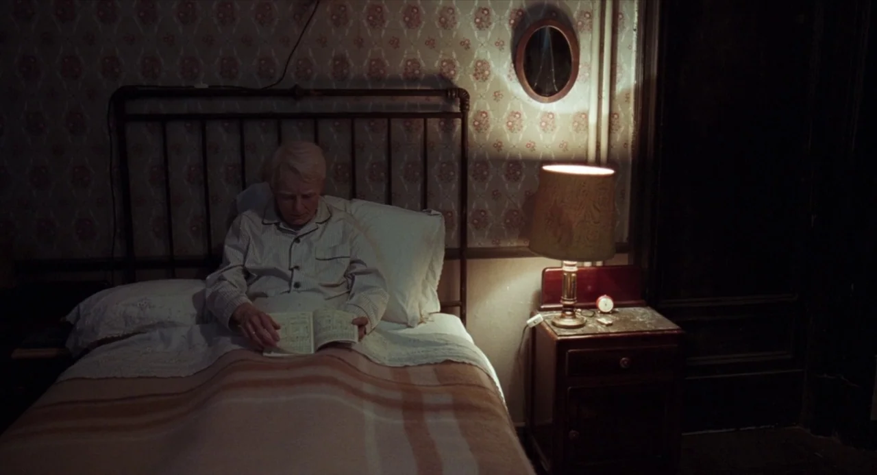 Suspiria (2018) movie still: medium — An interior bedroom with floral wallpaper and a metal-frame bed at night. An elderly man in pajamas sits…