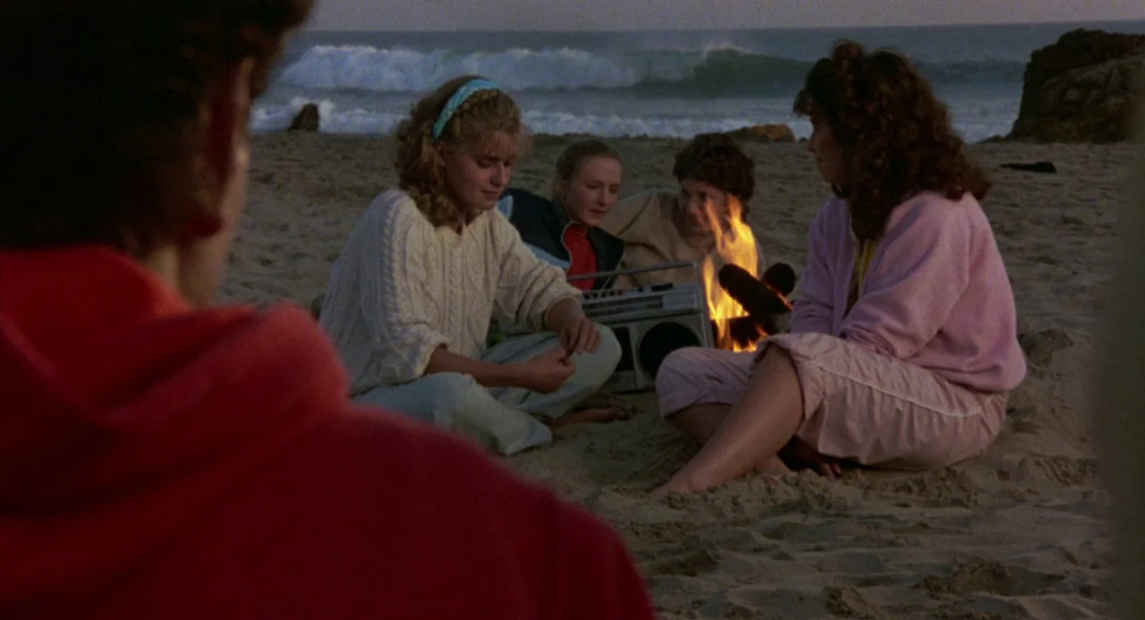The Karate Kid (1984) movie still: medium — A group of teenagers sits on a sandy beach at dusk near a campfire. They are gathered around a portable…