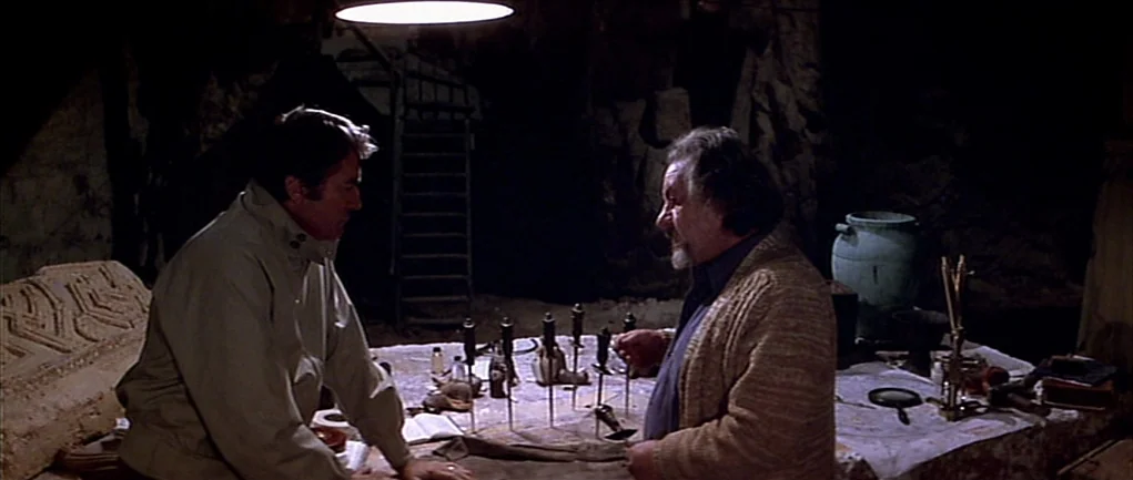 The Omen (1976) movie still: medium — An underground stone enclosure contains a table covered with archaeological tools and metal instruments. Two…