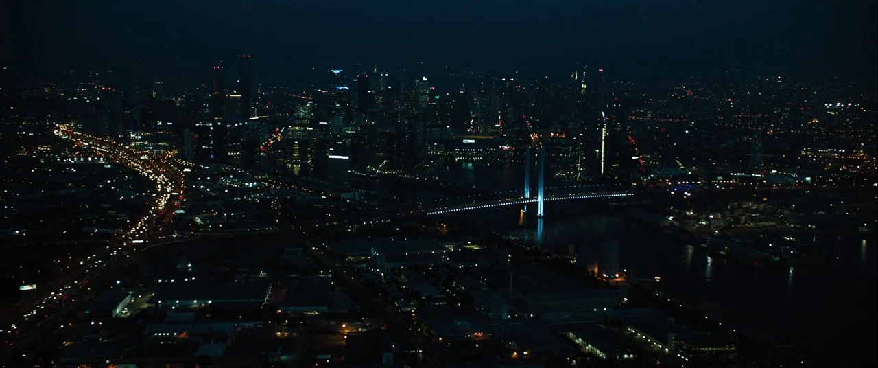 Upgrade (2018) movie still: establishing — A vast night aerial view of a sprawling city with a prominent river bridge. The horizon glows with…