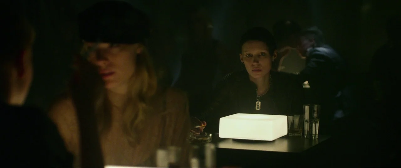 The Girl With The Dragon Tattoo (2011) movie still: medium — A dimly lit club with a low-light atmosphere features dark shadows. A woman sits at a…