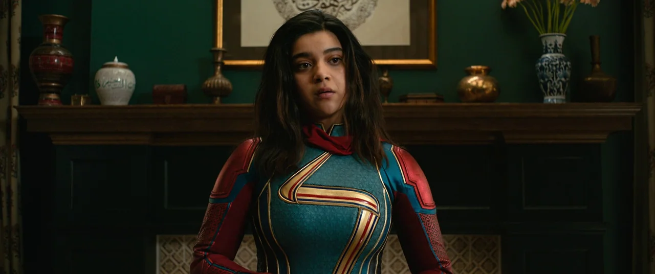 The Marvels (2023) movie still: medium — A dark green wall features a wooden mantle topped with vases and a framed print. A young woman in a…