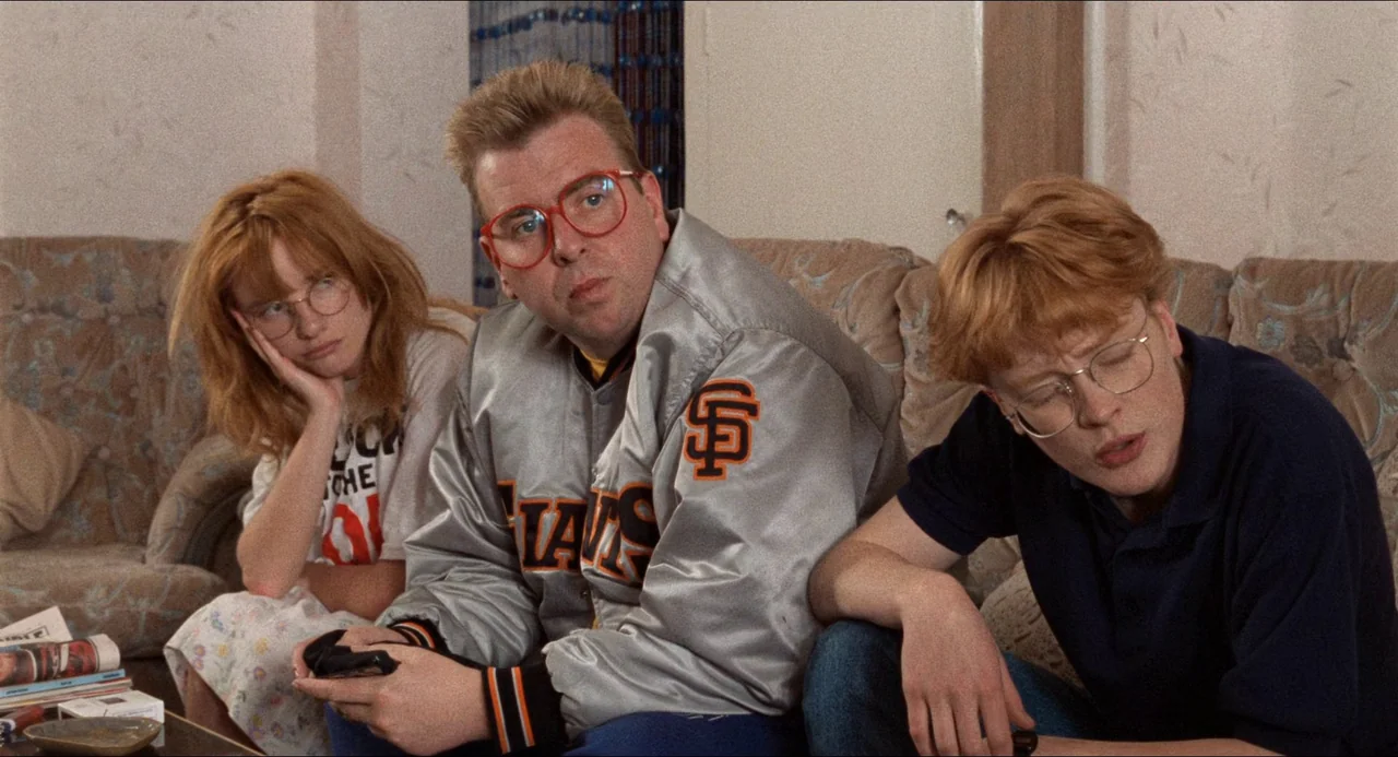 Life Is Sweet (1990) movie still: medium — Three people sit on a beige floral couch in a residential living room setting. The two men and one woman…