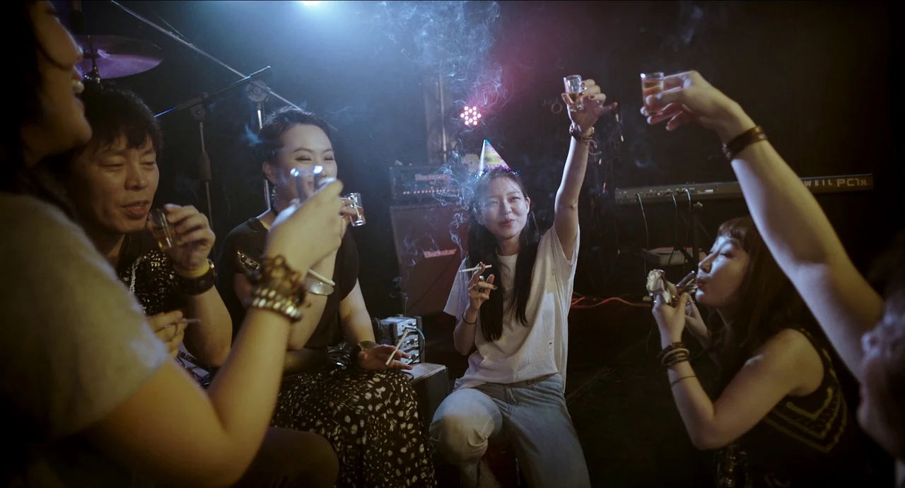 Microhabitat (2017) movie still: medium — A dimly lit backstage area of a music venue filled with haze and equipment. A group of friends sits on the…
