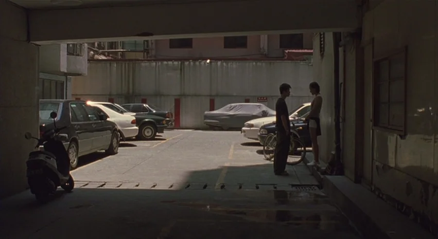 Yi-Yi (A One and A Two) (2000) movie still: wide — An asphalt parking area sits beneath a low concrete building overhang in daylight. A young man…