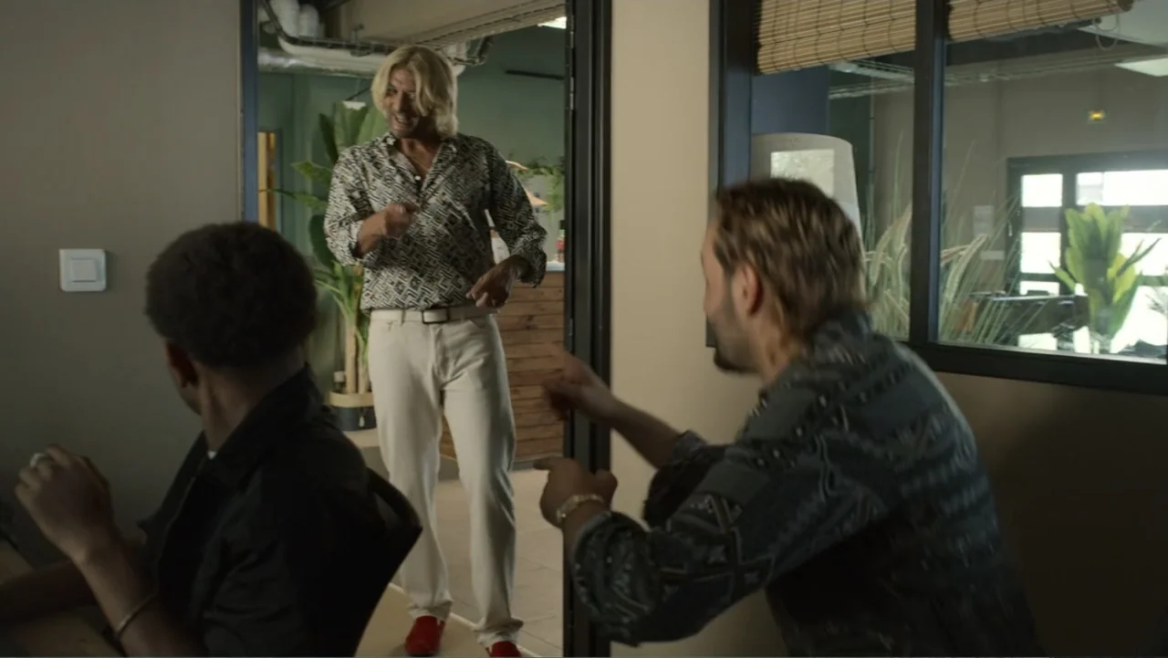 Unknown #1 movie still: medium — A modern office interior features glass partitions, potted plants, and beige walls. One man enters through a door…