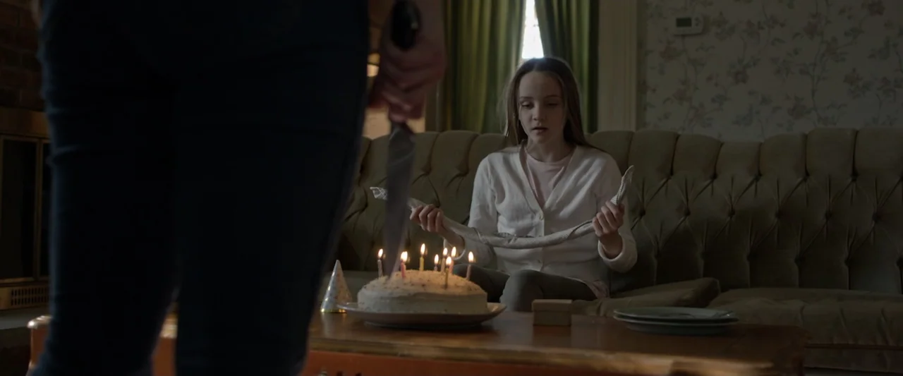 Motherly (2022) movie still: medium — A domestic living room contains a brown sofa and a birthday cake on a table. A person holds a sharp kitchen…