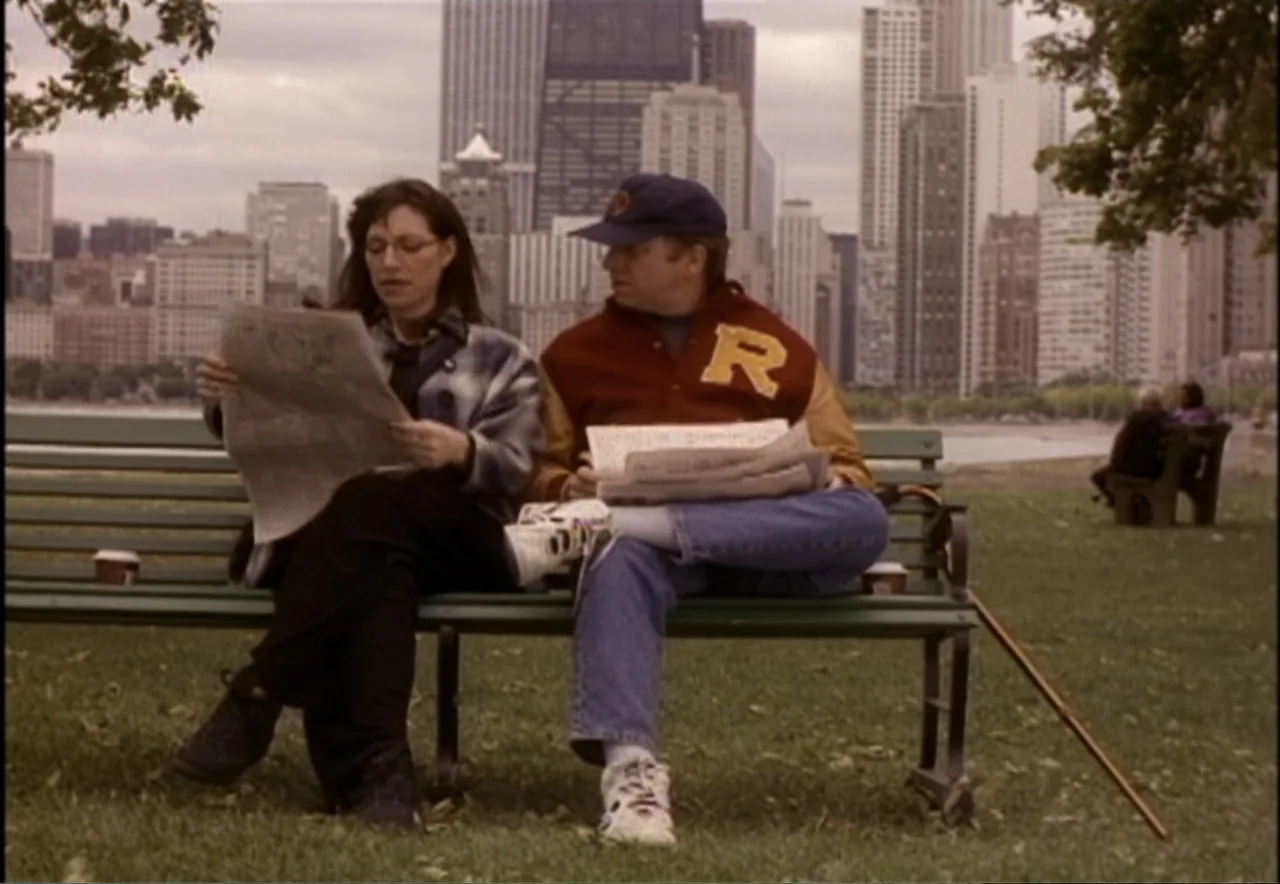 Chance of a Lifetime (1998) movie still: fullbody — A green wooden bench sits in a grassy park before a city skyline backdrop. A man and a woman sit…