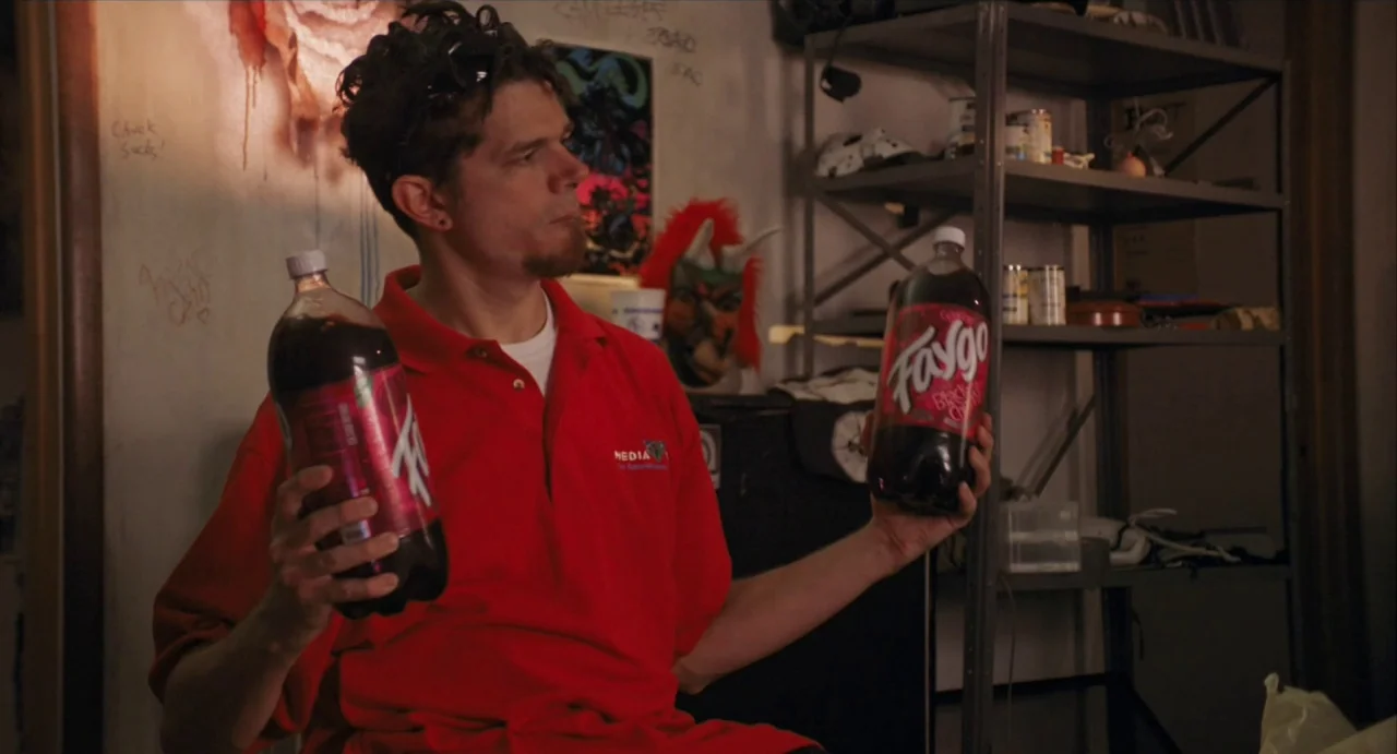 Relaxer (2018) movie still: medium — A cluttered room features a man holding two plastic soda bottles in his hands. He wears a red polo shirt while…