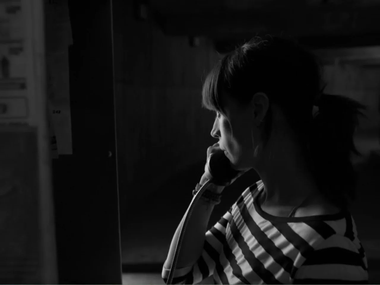 Güeros (2014) movie still: medium — A dimly lit telephone booth with minimal visible background architecture. A young woman in a striped shirt…