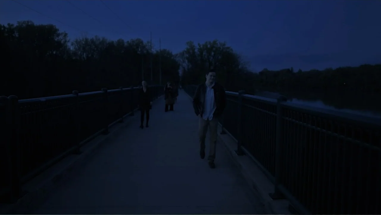 Ask Me to Dance (2022) movie still: fullbody — A dark pedestrian bridge spans over water surrounded by trees under a night sky. A man walks toward…