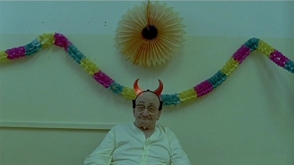 Import Export (2007) movie still: medium — An elderly man wearing red plastic devil horns sits against a plain wall. A paper fan and a colorful…